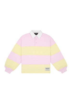 Young woman wearing a pastel pink and yellow striped long-sleeve polo shirt and blue checkered shorts, standing against a plain background.