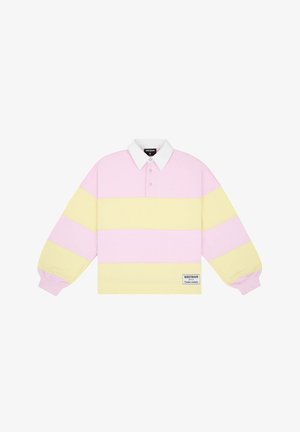 Młoda kobieta nosząca pastelowo różową i żółtą koszulkę polo z długim rękawem w paski oraz niebieskie szorty w kratę, stojąca na jednolitym tle.