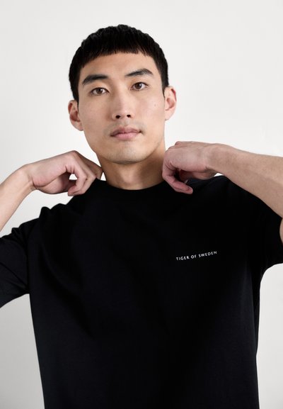 Must must-shirt lühikeste varrukatega, ümar kaelus, millel on rinnal valge tekst "TIGER OF SWEDEN". Sile kangatekstuur.