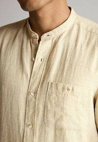 Chemise boutonnée beige clair avec un col mandarin, tissu texturé présentant un motif à grille subtil, et une poche poitrine unique.