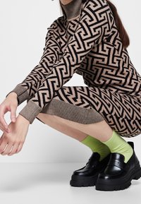 Abito a maglia con motivo geometrico marrone e nero, colletto alto, polsini a coste, abbinato a mocassini neri chunky e calzini verdi vivaci.