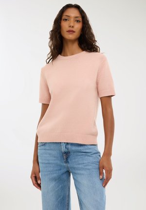 Maglione lavorato a maglia a maniche corte di colore rosa pastello con scollatura e orlo a coste. Abbinato a jeans blu chiaro, che mostrano una vestibilità rilassata e una trama morbida.