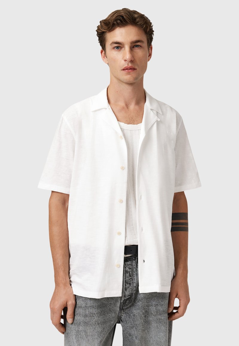 Chemise blanche à manches courtes en tissu léger avec un bouton sur le devant. Présente une coupe décontractée et une texture lisse, associée à un jean gris.