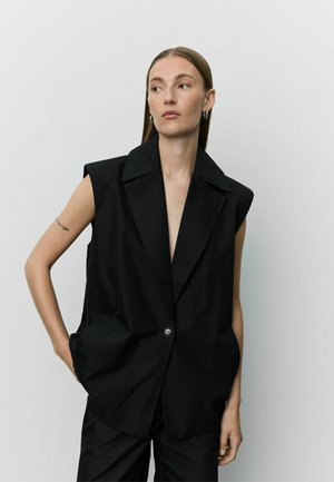 Donna con capelli castani e lisci indossa un blazer nero senza maniche con un bottone, in piedi con una mano in tasca davanti a uno sfondo semplice.