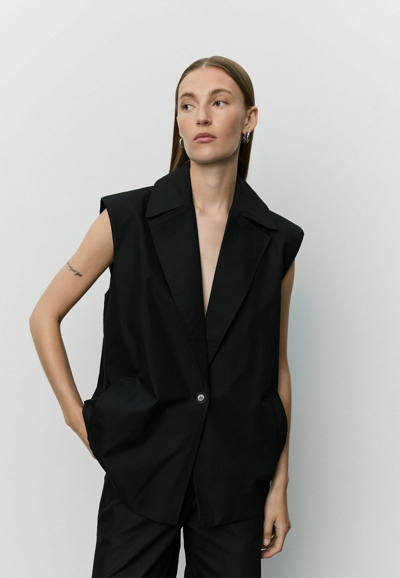 Femme aux cheveux bruns et raides portant un blazer noir sans manches avec un bouton, debout une main dans la poche devant un fond uni.