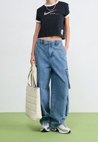 Černé zkrácené tričko s bílým límečkem a červeným logem, modré džínové cargo kalhoty s bočními kapsami, světle šedé tenisky a béžová taška.