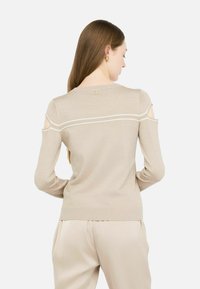 Beiger Pullover mit offenem Schulterdesign, geripptem Halsauschnitt und Bündchen sowie einem dezenten Glanz. Verfügt über einen kleinen goldenen Akzent auf der Rückseite.