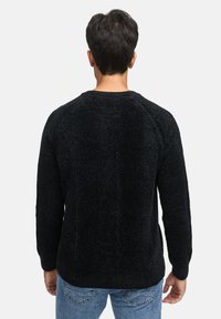 Uomo con i capelli corti e scuri, che indossa un maglione nero a trama e jeans blu, in piedi di spalle contro uno sfondo chiaro e semplice.