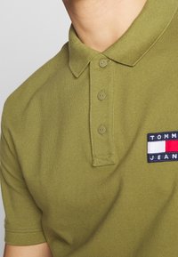 Olivově zelené polo tričko se třemi knoflíky a obdélníkovou nášivkou loga Tommy Jeans na hrudi.