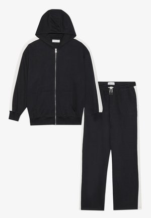 Chaqueta con capucha negra y cremallera, con rayas laterales blancas y pantalones de chándal negros a juego que presentan detalles de rayas blancas. Tejido suave, diseño casual.