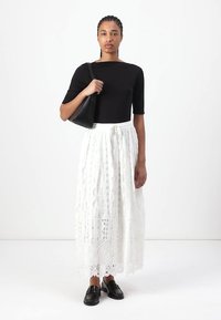 Zwarte top met korte mouwen, witte maxi rok van kant met ingewikkelde patronen, elastische taille, en zwarte instap schoenen. Model draagt een zwarte handtas.