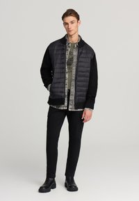 Giacca nera imbottita con fondo in maglia nera, sovrapposta a una camicia grigia con motivo, abbinata a pantaloni cargo neri e stivali neri robusti.