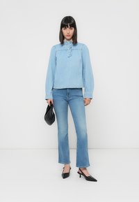 Woman standing wearing light blue ruffled neck blouse, medium wash jeans, black pointed heels, holding small black handbag.