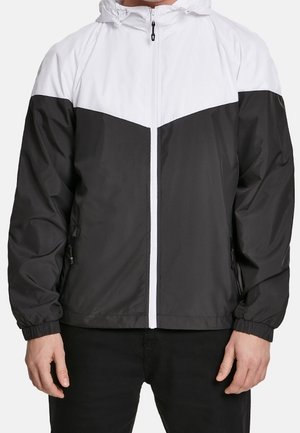 Homme portant une veste coupe-vent noire et blanche à fermeture éclair, avec des poignets élastiques et des poches latérales zippées.