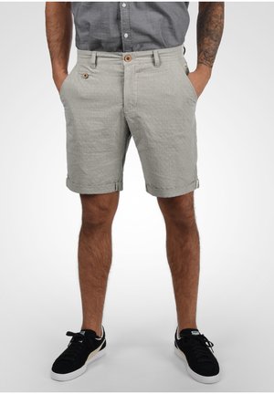 Mann trägt hellgraue Shorts mit umgeschlagenem Saum, graues Hemd, schwarz-weiße Turnschuhe, Hände in den Taschen und sichtbarer tätowierter rechter Arm.