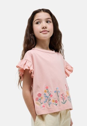 Jeune fille aux longs cheveux bruns portant un haut rose à manches volantées avec des fleurs colorées brodées le long de l'ourlet, un pantalon beige, les mains dans les poches.