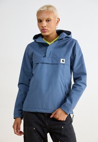 Giacca blu con cappuccio, dotata di tasca frontale con zip e design a pullover. Realizzata in materiale leggero, presenta una patch con logo e polsini regolabili.