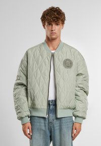 Giacca bomber trapuntata verde chiaro con polsini e cintura a coste; presenta un patch rotondo con logo sul lato sinistro del petto e chiusura con zip davanti.