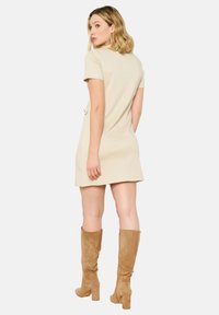 Beige Kurzarmkleid mit geradem Schnitt und zwei seitlichen Taschen. Kombiniert mit kniehohen, taupefarbenen Wildlederstiefeln und einer glatten Textur.