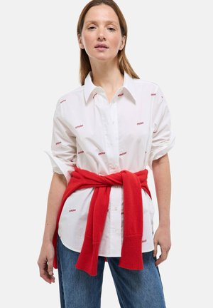 Femme portant une chemise blanche à boutons avec un motif de texte rouge, un pull rouge noué à la taille, et un jean bleu, debout devant un fond blanc.