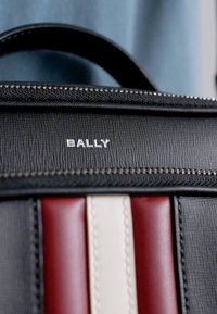 Geantă din piele texturată neagră cu dungi orizontale roșii și crem, având accesorii din metal argintiu și închidere cu fermoar. Logo-ul "BALLY" este vizibil.