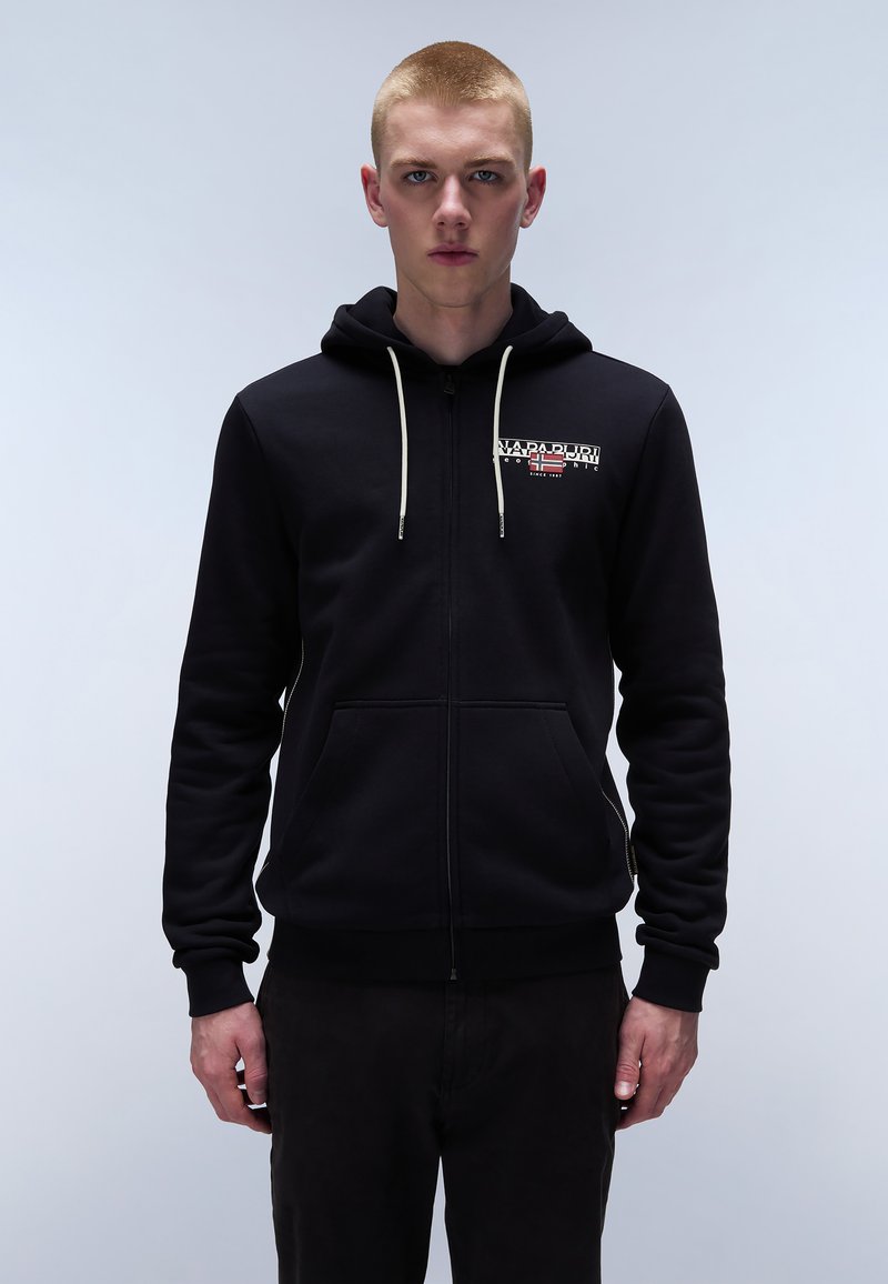 Sudadera con cremallera negra hecha de algodón grueso, con un logotipo en el frente, capucha con cordón y bolsillos laterales. El modelo está de pie contra un fondo liso.