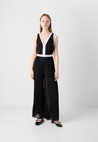 Jumpsuit plissé noir avec jambes larges, décolleté en V et une ceinture d'accent blanc. Le modèle porte des chaussures plates noires, avec une texture lisse.