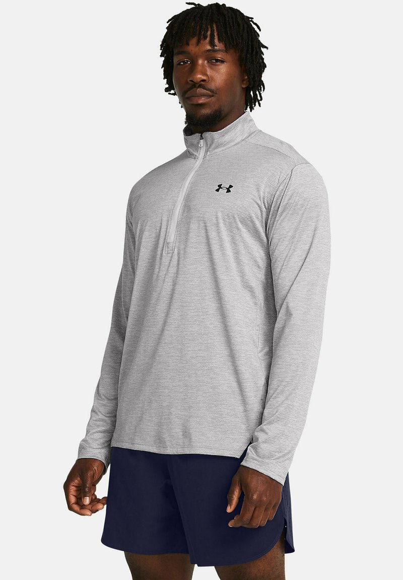 Grau melierter Pullover mit langen Ärmeln und halbem Reißverschluss. Ausgestattet mit einem strukturierten Stoff und einem kleinen schwarzen Logo auf der Brust. Kombiniert mit marineblauen Shorts.