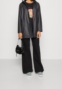 Veste noire imperméable, pantalon noir ample, et baskets vertes à motif damier. Petit sac à main noir avec détail chaîne. T-shirt graphique en dessous.