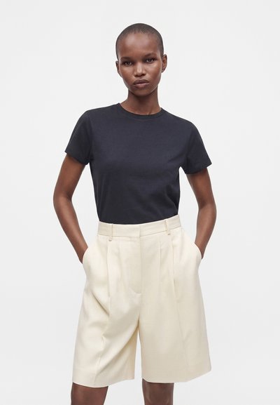 Femme aux cheveux courts portant un t-shirt noir à manches courtes et un short beige ample taille haute, les mains dans les poches.
