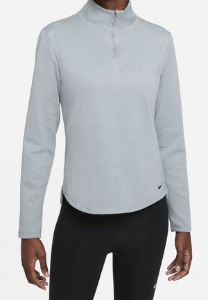 Person trägt ein hellgraues Langarm-Quarter-Zip-Oberteil und schwarze Leggings, steht vor einem schlichten weißen Hintergrund.