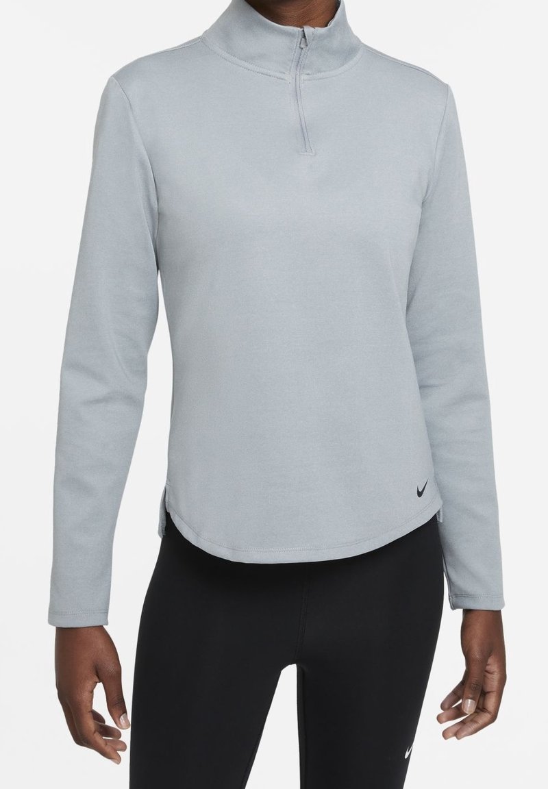 Osoba w szarej, jasnej bluzie z długim rękawem i suwakiem do połowy oraz czarnych legginsach, stojąca na tle jednolitego, białego tła.