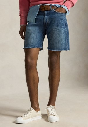 Férfi, kék farmer rövidnadrágban, nyers szegéllyel, fehér tornacipővel, barna övvel és rózsaszín pulóverrel, kék ing fölött.