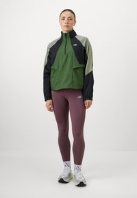 Veste coupe-vent en vert, noir et gris clair ; possède un col haut, une fermeture éclair dissimulée et une poche avant. Portée avec des leggings violet et des baskets.
