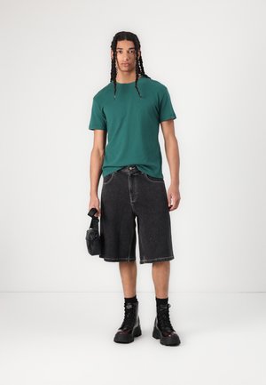 Grønn bomull t-skjorte, mørke denimshorts, svarte militærstøvler. Modellen holder en liten svart veske. Enkel design; avslappet passform.