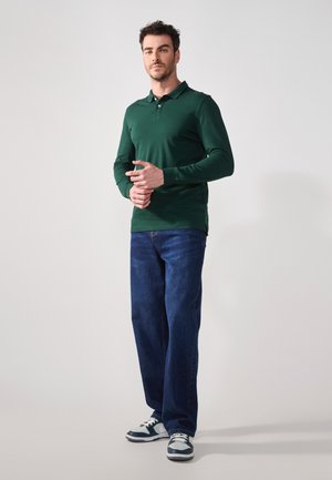 Žalias polo marškinėliai ilgomis rankovėmis, su aptemptu siluetu, šoninėmis skeltuvėmis, derinami su laisvais mėlynais džinsais ir sportbačiais su baltais ir tamsiais akcentais.
