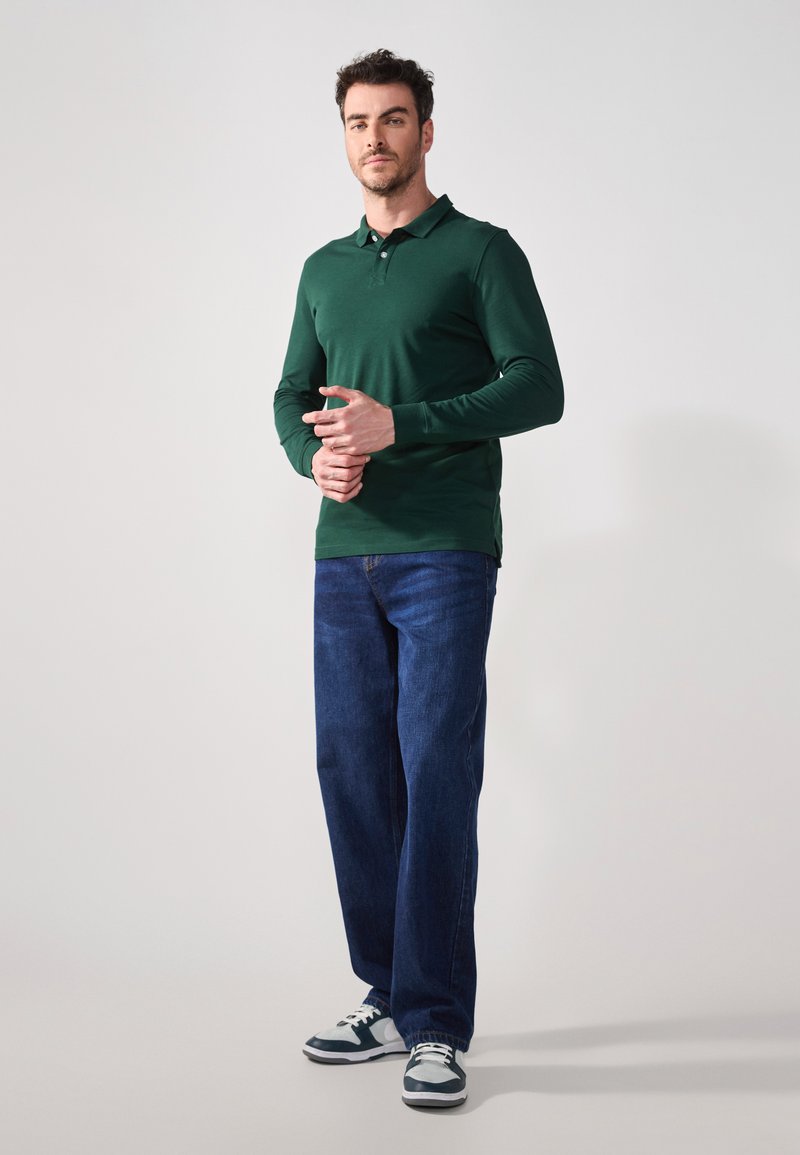 Grön långärmad polo skjorta i tapered fit, med sidsnitt, kombinerad med löst sittande blå jeans och sneakers med vita och mörka accenter.
