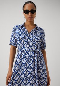 Blue and white collared dress with a leaf pattern, short sleeves, tie belt at the waist, made of lightweight fabric. Black sunglasses worn.
