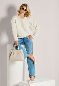 Vrouw in een gebreide crèmekleurige trui, blauwe jeans, crèmekleurige hakken, met een crèmekleurige handtas, die zonnebril draagt, poseert tegen een lichte muur op witte blokken.