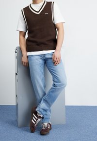 Chaleco marrón de punto con ribete blanco sobre una camiseta blanca, combinado con vaqueros azul claro y zapatillas marrones con rayas blancas.