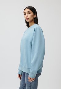 Felpa azzurra con maniche lunghe, scollo rotondo, texture morbida e un logo bianco minimale sul davanti. Abbinata a jeans blu.