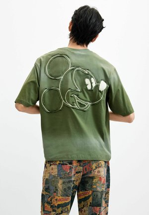 Hombre que lleva una camiseta verde oliva con una gran cara de Mickey Mouse cosida en la espalda, combinada con pantalones multicolores con estampados de estilo cómic.