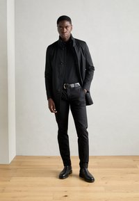 Zwarte trenchcoat over een zwarte coltrui, gecombineerd met zwarte broek en enkellaarsjes. Gladde textuur, slim fit en minimalistisch design.