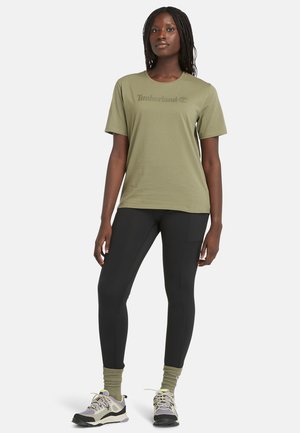 Olijfgroene katoenen T-shirt met korte mouwen en het "Timberland" logo; gecombineerd met zwarte leggings en grijze wandelschoenen.