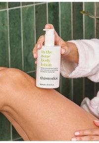 Weißer Kunststoffpumpspender mit der Beschriftung "In the Zone Body Lotion" in grüner Schrift, vor einem grünen gefliesten Hintergrund gehalten in einer Hand.