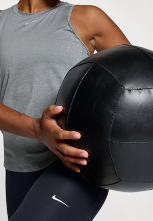 Personne en débardeur gris et leggings noirs tenant un grand ballon médicinal noir sur fond blanc.