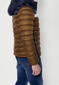 Homme portant une veste matelassée marron et bleu marine avec une capuche et un jean bleu, debout devant un fond clair uni.