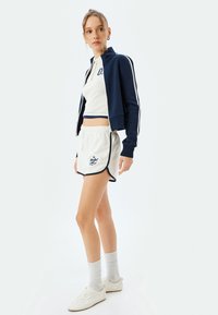 Giacca blu navy con strisce bianche, top corto bianco, pantaloni corti beige con bordi blu e testo, abbinati a scarpe bianche e calzini grigi.