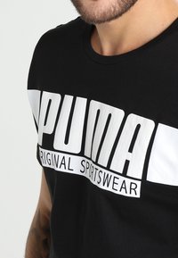 Muž oblečený v černém tričku s velkým bílým logem "PUMA" a textem "ORIGINAL SPORTSWEAR" přes hruď na jednoduchém pozadí.