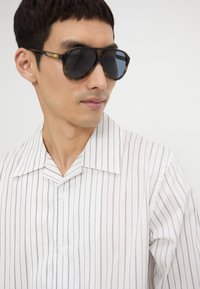 Camisa blanca con finas rayas verticales en navy, que presenta un amplio collar y cierre de botones. Gafas de sol oscuras con un grueso marco de carey.